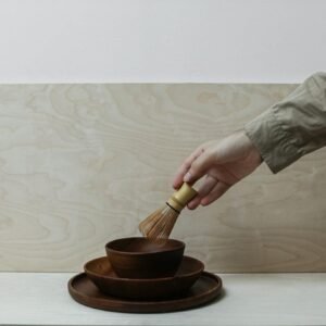 A hand holds a bamboo whisk over stacked wooden bowls, set against a minimalist background.