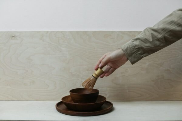 A hand holds a bamboo whisk over stacked wooden bowls, set against a minimalist background.